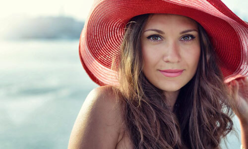 Portrait picture of a beautiful woman, happy with her face lift with neck lift procedure in San Jose, Costa Rica.  The woman has long dark hair, is looking directly into the camera and is wearing a soft red beach hat highlighting her face lift with neck lift.